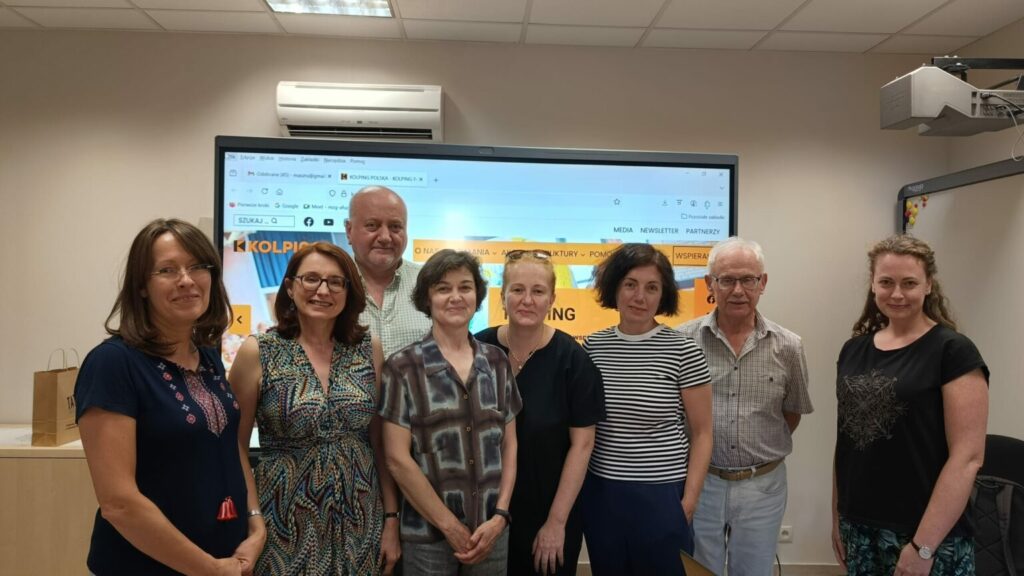 Grupa dorosłych osób stoi uśmiechnięta i pozuje do zdjecia. W tle tablica interaktywna z logotypem "KOLPING".