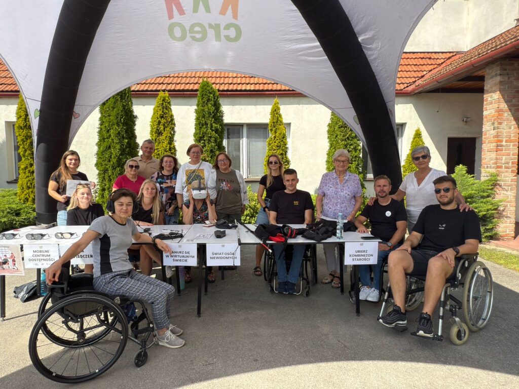 zdjecie przedstawia grupę osób stojących lub siedzących w namiocie wystawienniczym. dwie osoby siedzą na wózkach inwalidzkich.