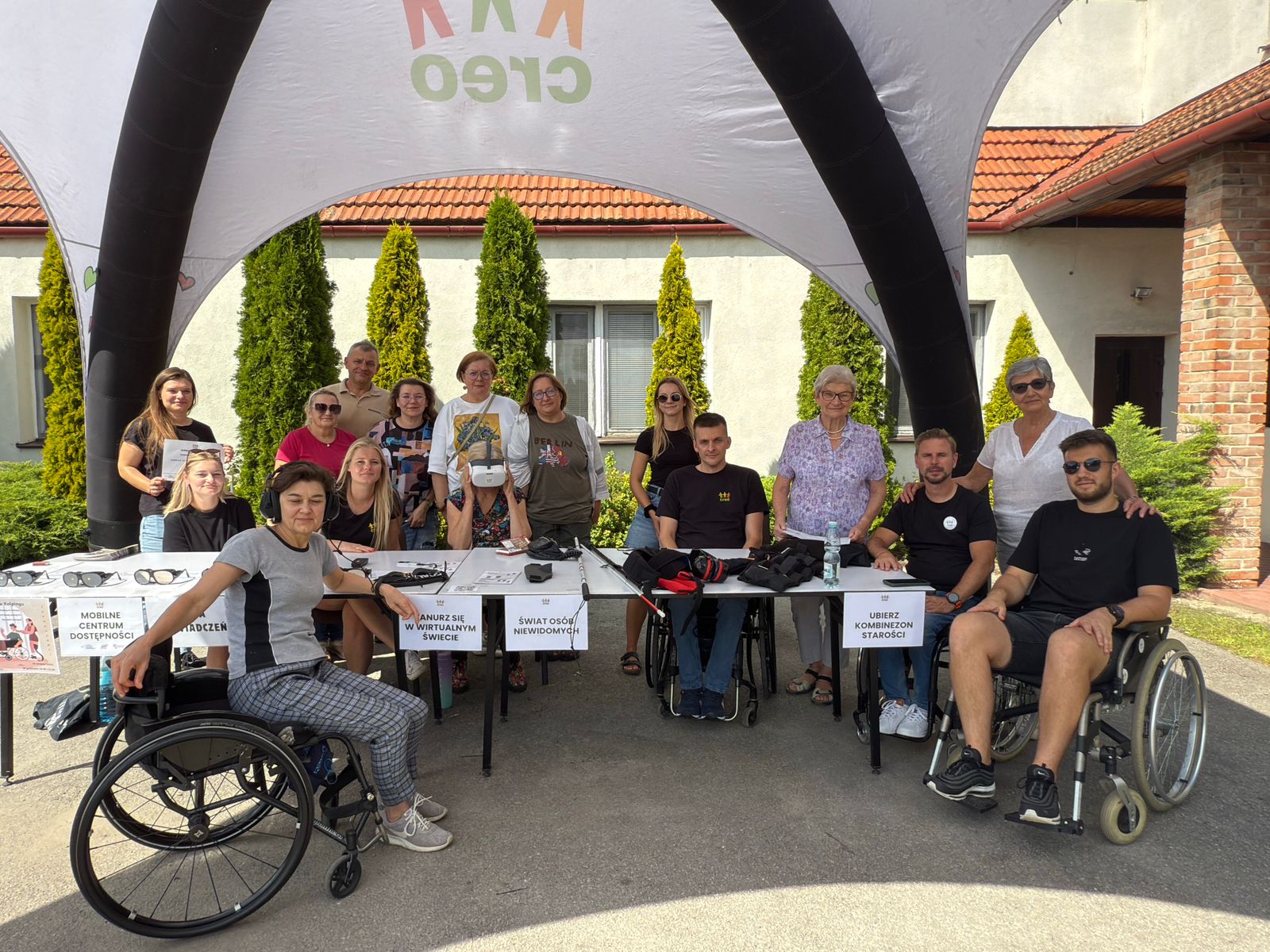 zdjecie przedstawia grupę osób stojących lub siedzących w namiocie wystawienniczym. dwie osoby siedzą na wózkach inwalidzkich.