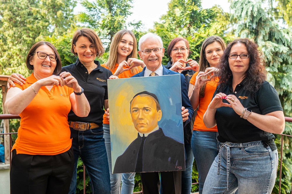 Grupa osób w koszulkach z logiem kolping pozuje do zdjęcia, trzymają obraz przedstawiający Adolpha Kolpinga. Ręce układają w kształt serduszek