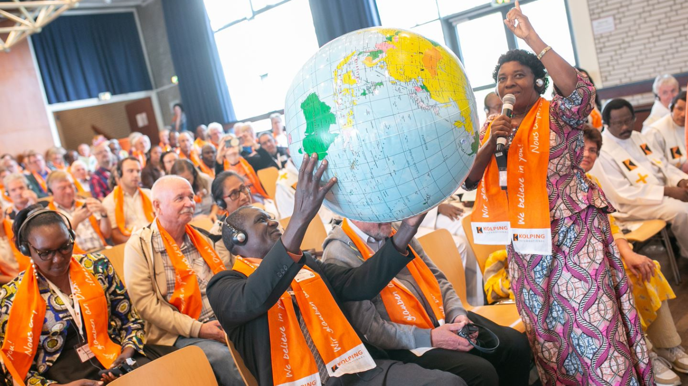 Uczestnicy siedzący w rzędach w trakcie dużego spotkania. Jeden z uczestników trzyma nadmuchany globus. Wszyscy uczestnicy mają pomarańczowe szaliki z logiem kolping
