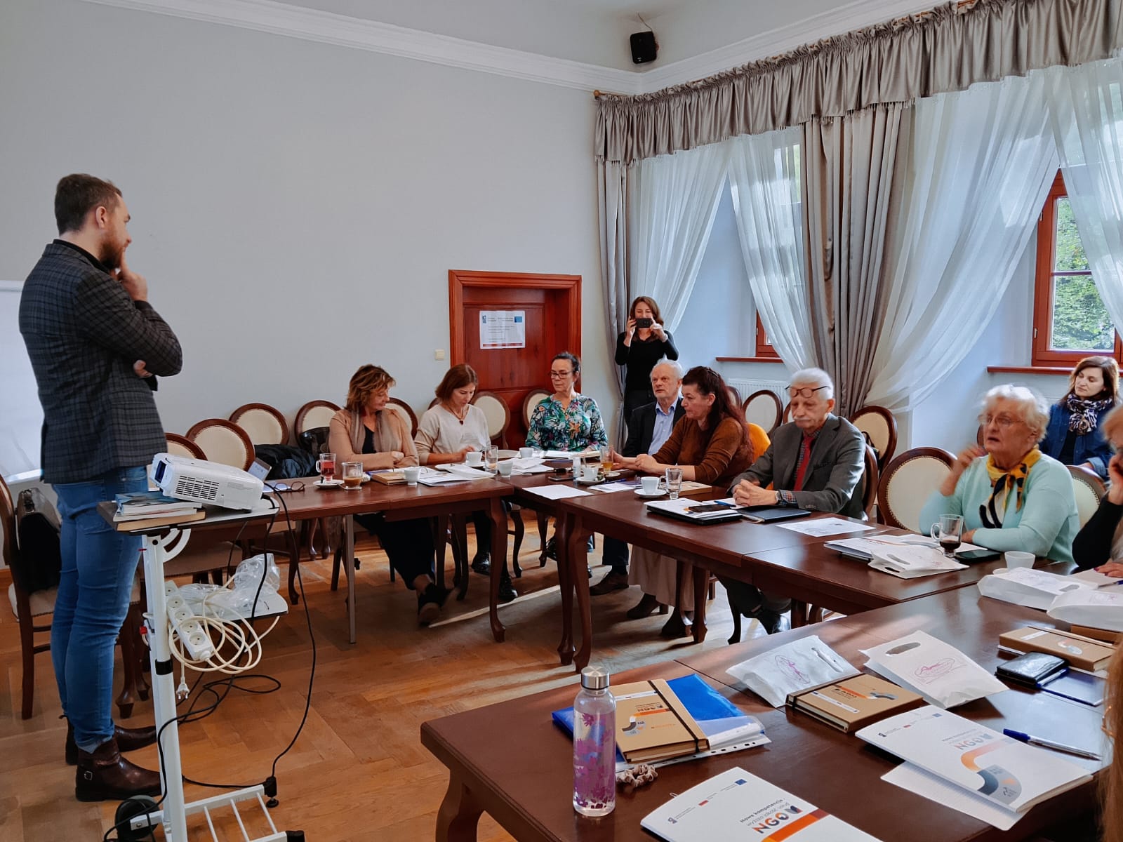 zdjecie przedstawia osoby siedzace przy stole sali szkoleniowej i uczestniczące w szkoleniu oraz trenera stojącego naprzeciwko nich