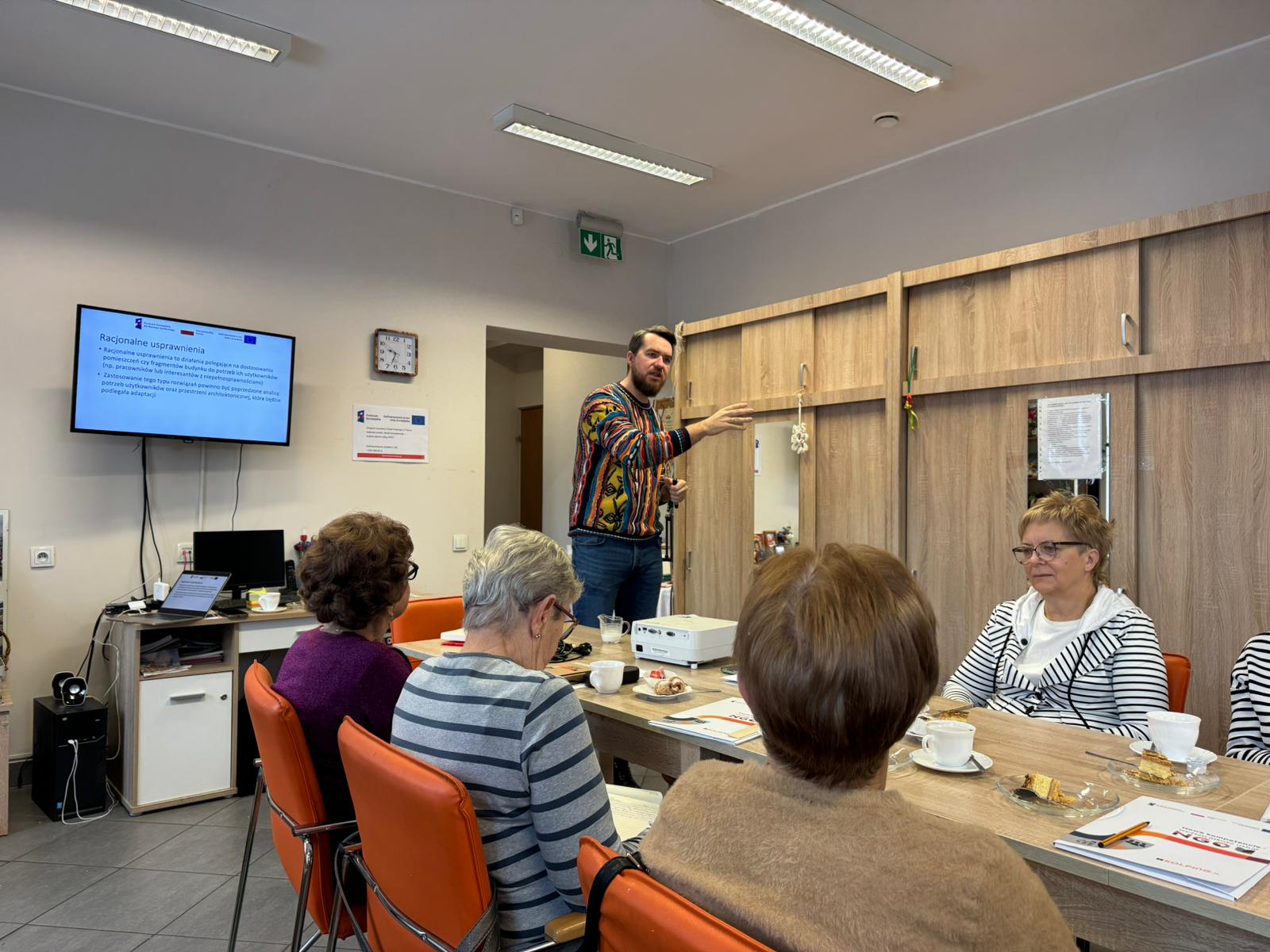 zdjęcie przedstawia grupę uczestników szkolenia, którzy siedzą przy stole i słuchają prowadzącego, który stoi w głębi sali obok ekranu z prezentacją.