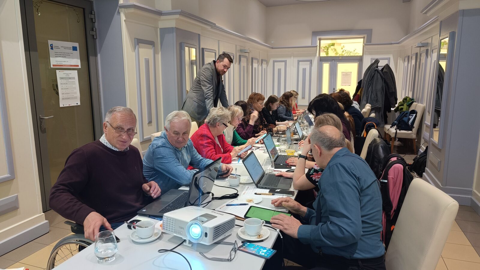 grupa uczestników szkolenia siedzi przy stołach z laptopami, trener stoi i sprawdza ich pracę