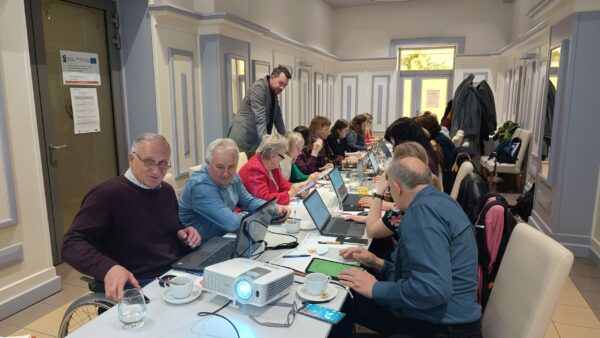 grupa uczestników szkolenia siedzi przy stołach z laptopami, trener stoi i sprawdza ich pracę