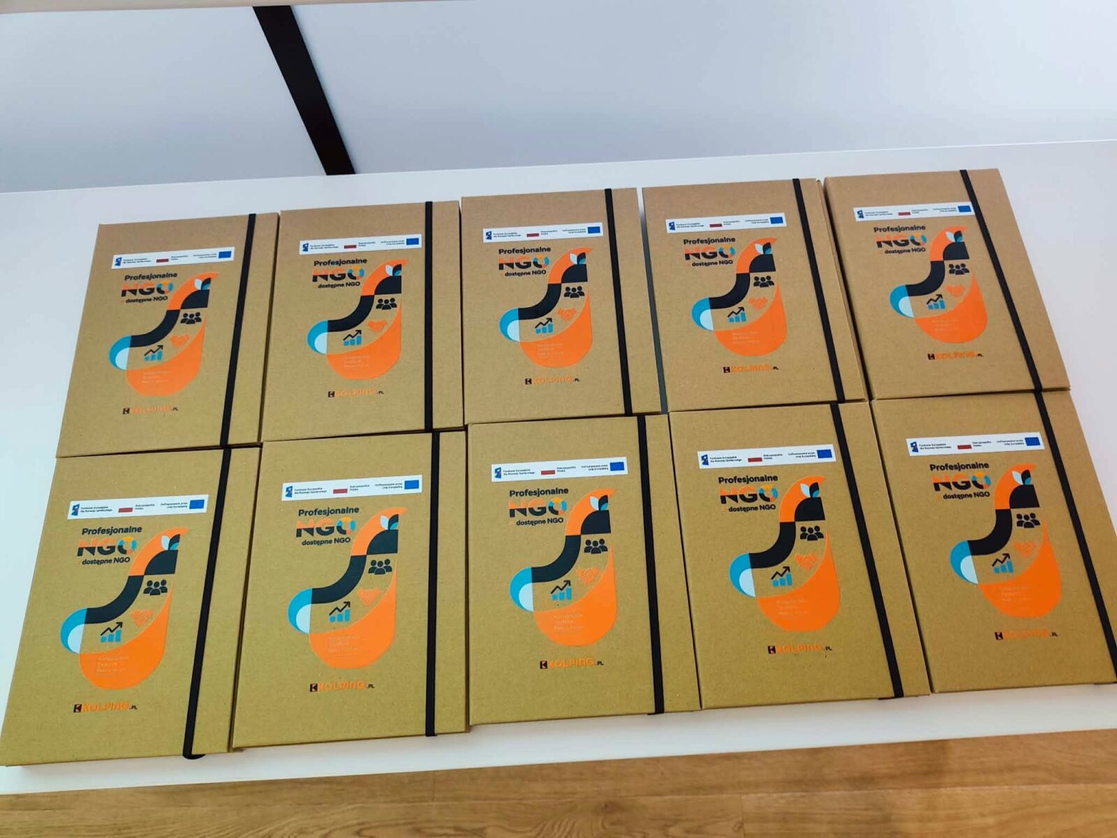 Na białym stole ułożono dziesięć egzemplarzy materiałów szkoleniowych w brązowych okładkach z kolorową grafiką. Na okładkach widnieje tytuł „Profesjonalne NGO = dostępne NGO” oraz logotypy projektu i instytucji finansujących. Materiały są przygotowane na potrzeby szkoleń realizowanych w ramach projektu.