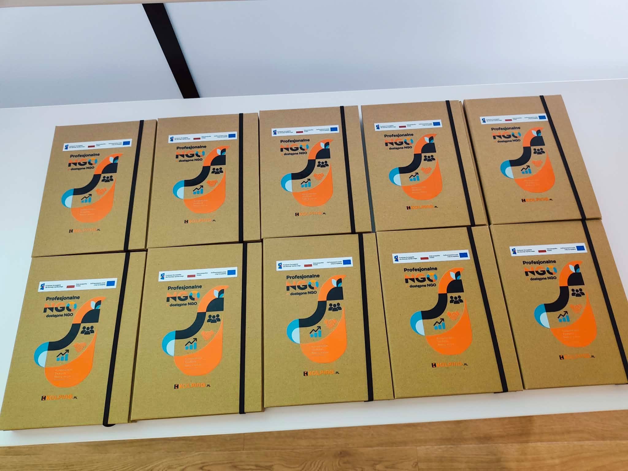 Na białym stole ułożono dziesięć egzemplarzy materiałów szkoleniowych w brązowych okładkach z kolorową grafiką. Na okładkach widnieje tytuł „Profesjonalne NGO = dostępne NGO” oraz logotypy projektu i instytucji finansujących. Materiały są przygotowane na potrzeby szkoleń realizowanych w ramach projektu.